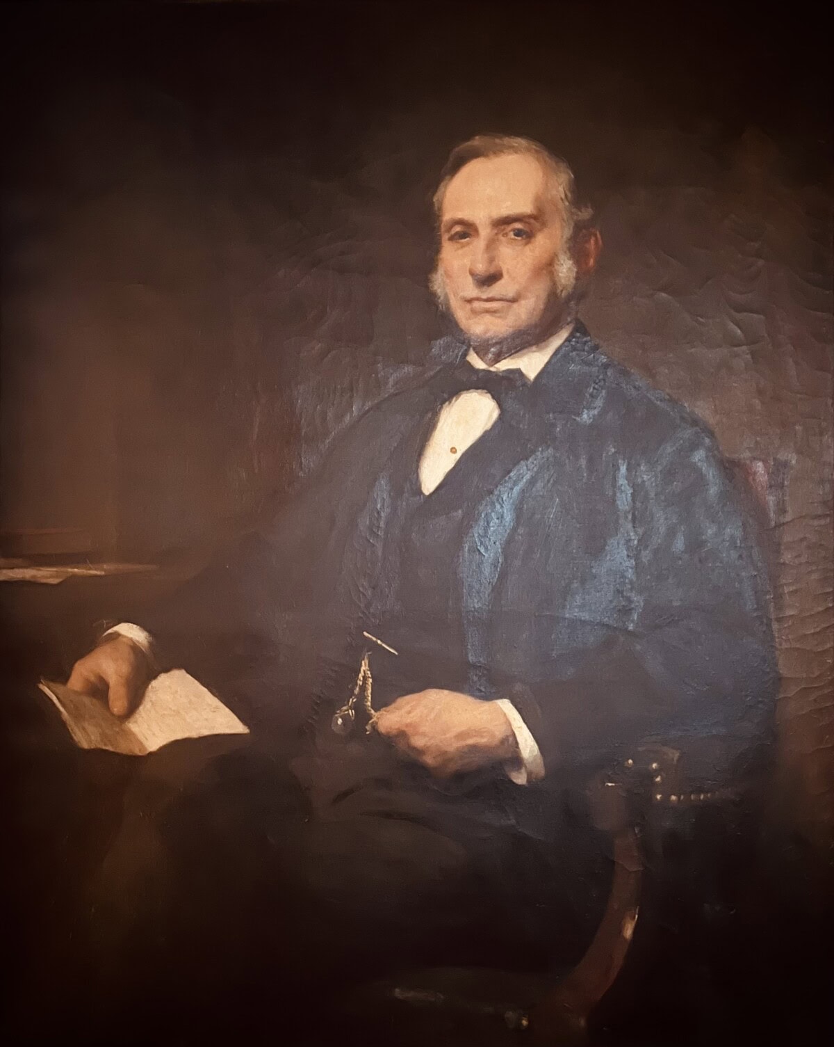 A formal oil portrait of an older man sitting in a chair, wearing a dark suit with a bow tie and blue vest, holding a paper in one hand and a pocket watch chain visible at his waist. The background is dark and muted.