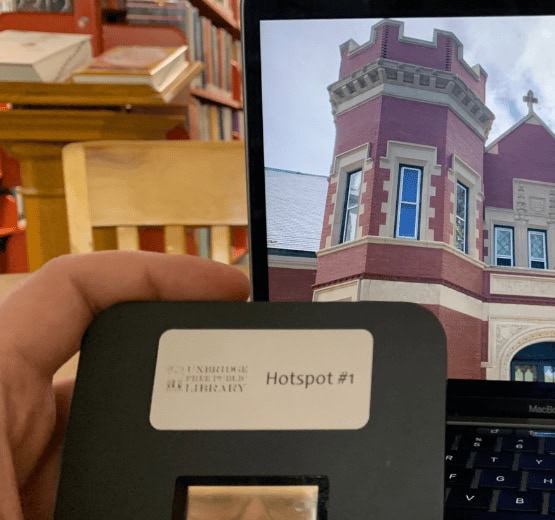 A hand holds a Wi-Fi hotspot device labeled Hotspot #1 in front of a laptop displaying an image of a red brick building with white trim and a tower. Bookshelves are visible in the background.