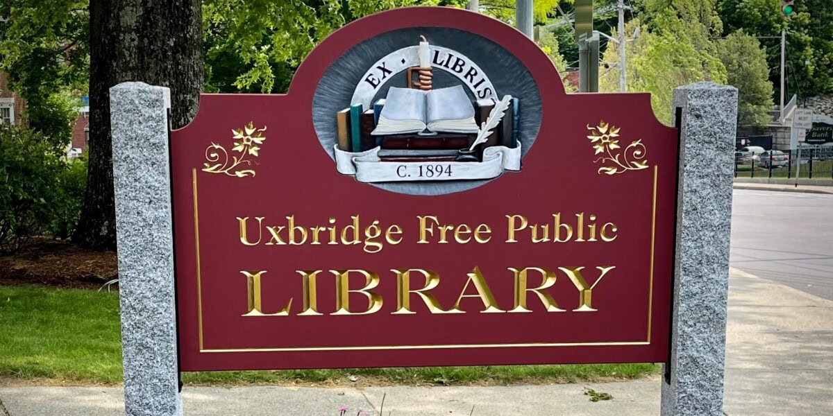 A maroon sign with gold letters reads Uxbridge Free Public Library and features an illustration of books and an open book with Ex Libris C. 1894 above it, flanked by two stone posts. Trees and a street are in the background.