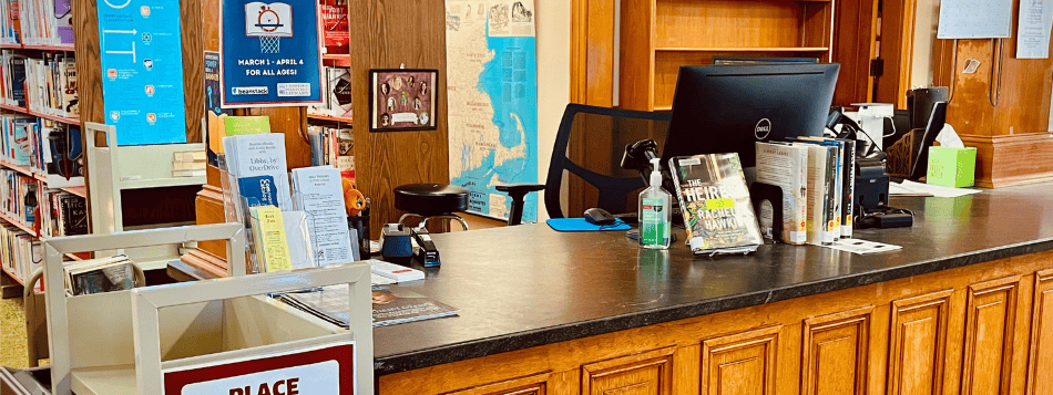 A wooden library checkout desk with a computer, books on display, hand sanitizer, and various flyers. Bookshelves filled with books and informational signs are visible in the background.