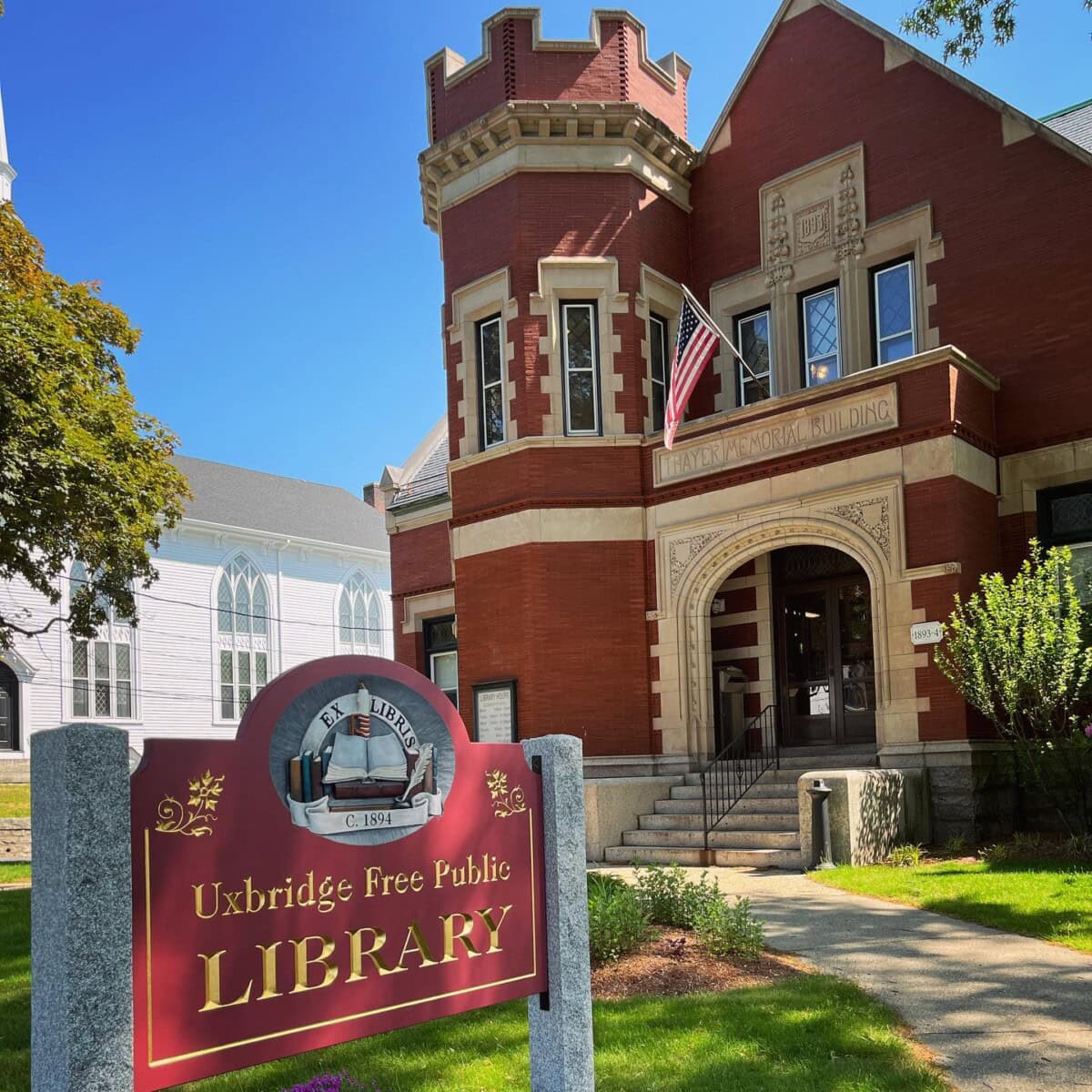 A red-brick building with a turret houses the Uxbridge Free Public Library. An American flag hangs by the entrance. A sign in front reads “Uxbridge Free Public Library.” Trees and a white building are in the background.