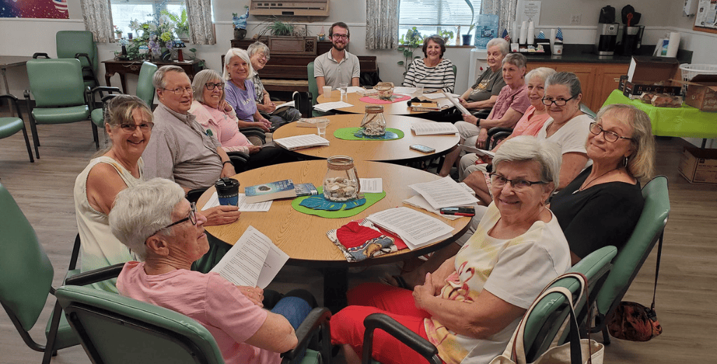 A group of seniors and one younger man sit around a large circular table in a bright community room, smiling at the camera. Papers, books, and decorations are on the table. Plants and a piano are in the background.